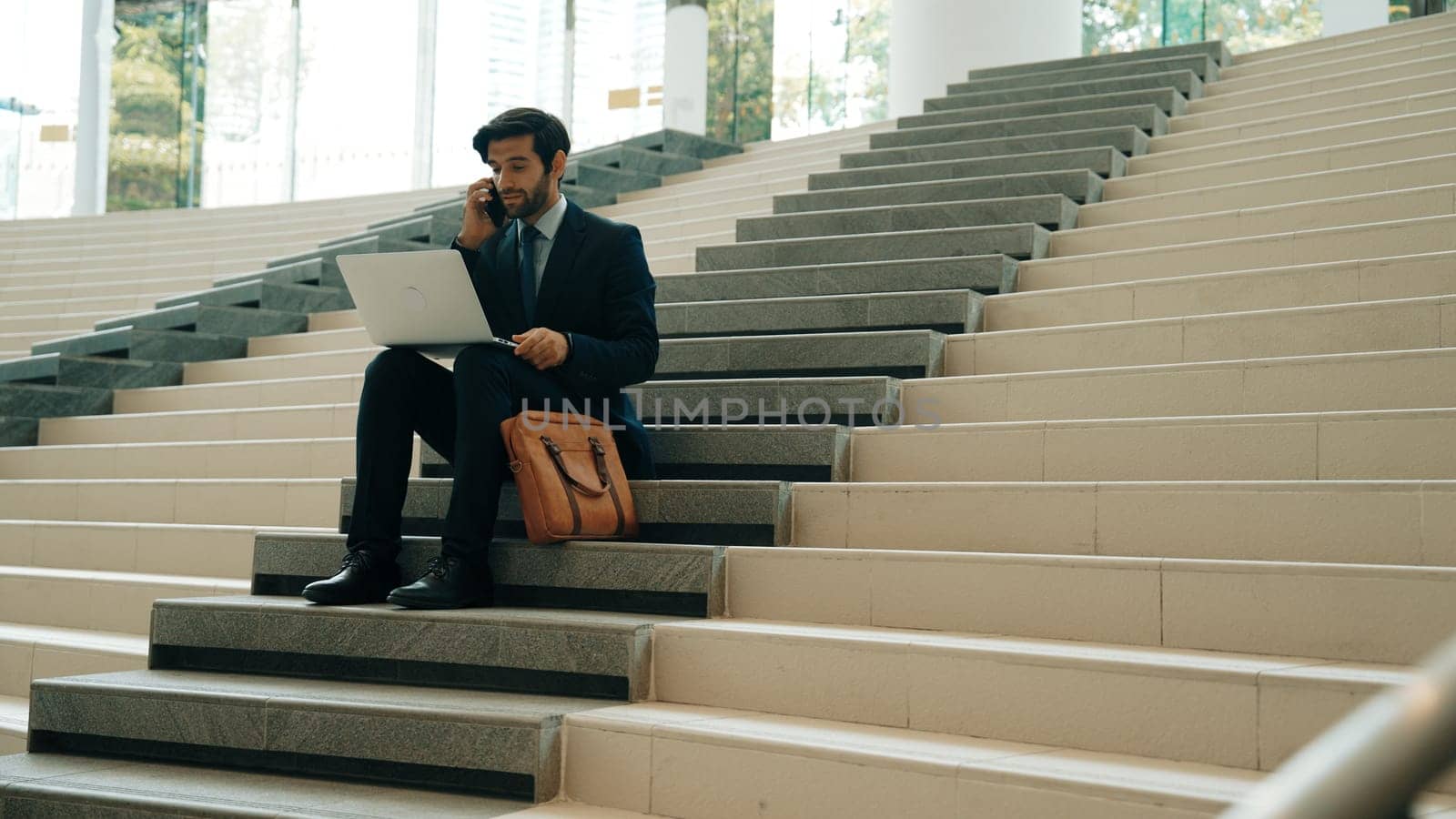 Skilled investor working or planing strategy by using laptop at stair. Professional business man wearing suit while working and typing data analysis by using laptop and talking to phone. Exultant.