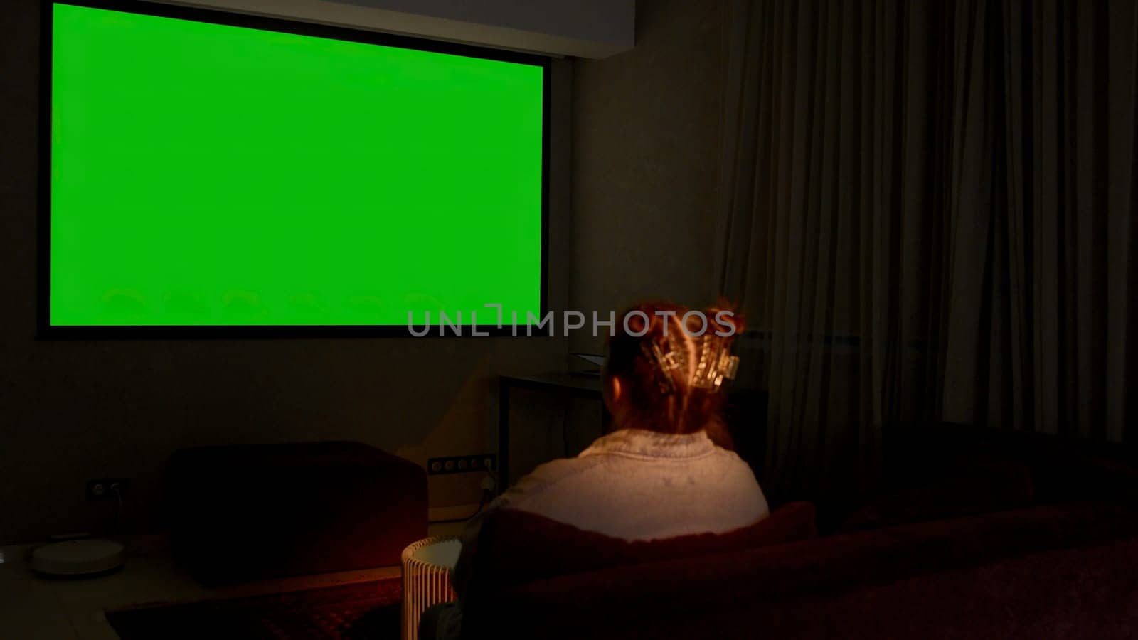 Woman watching television in living room late in the evening. Media. Young girl sitting on sofa against tv with chroma key green screen, back view