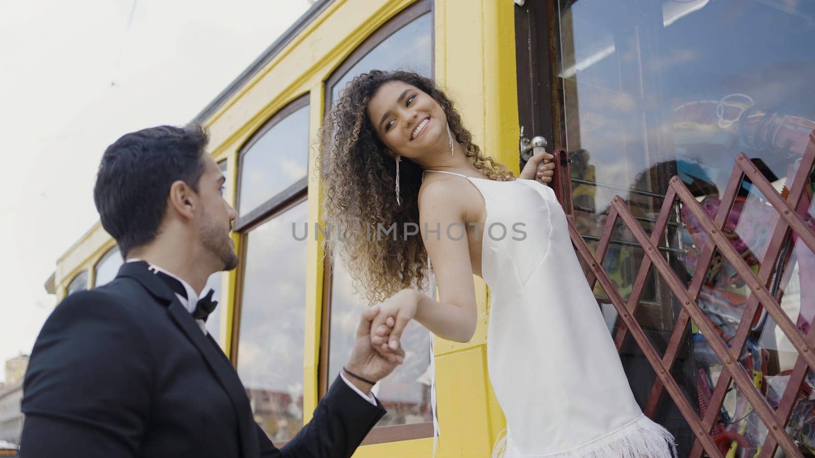Romantic date outdoors in the city street. Action. Woman on the stairs of the tram car with her hand in a hand of a man in black suit. by Mediawhalestock