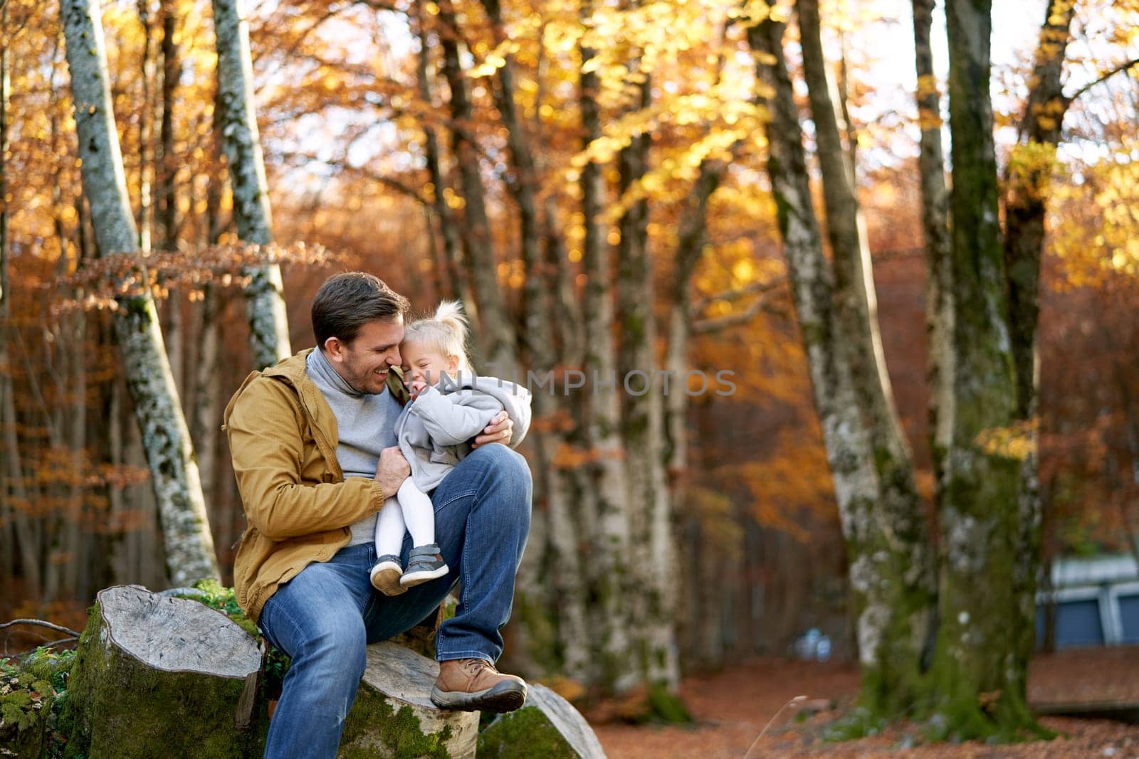Dad hugging a smiling little daughter sitting on his lap on a stump in the forest by Nadtochiy