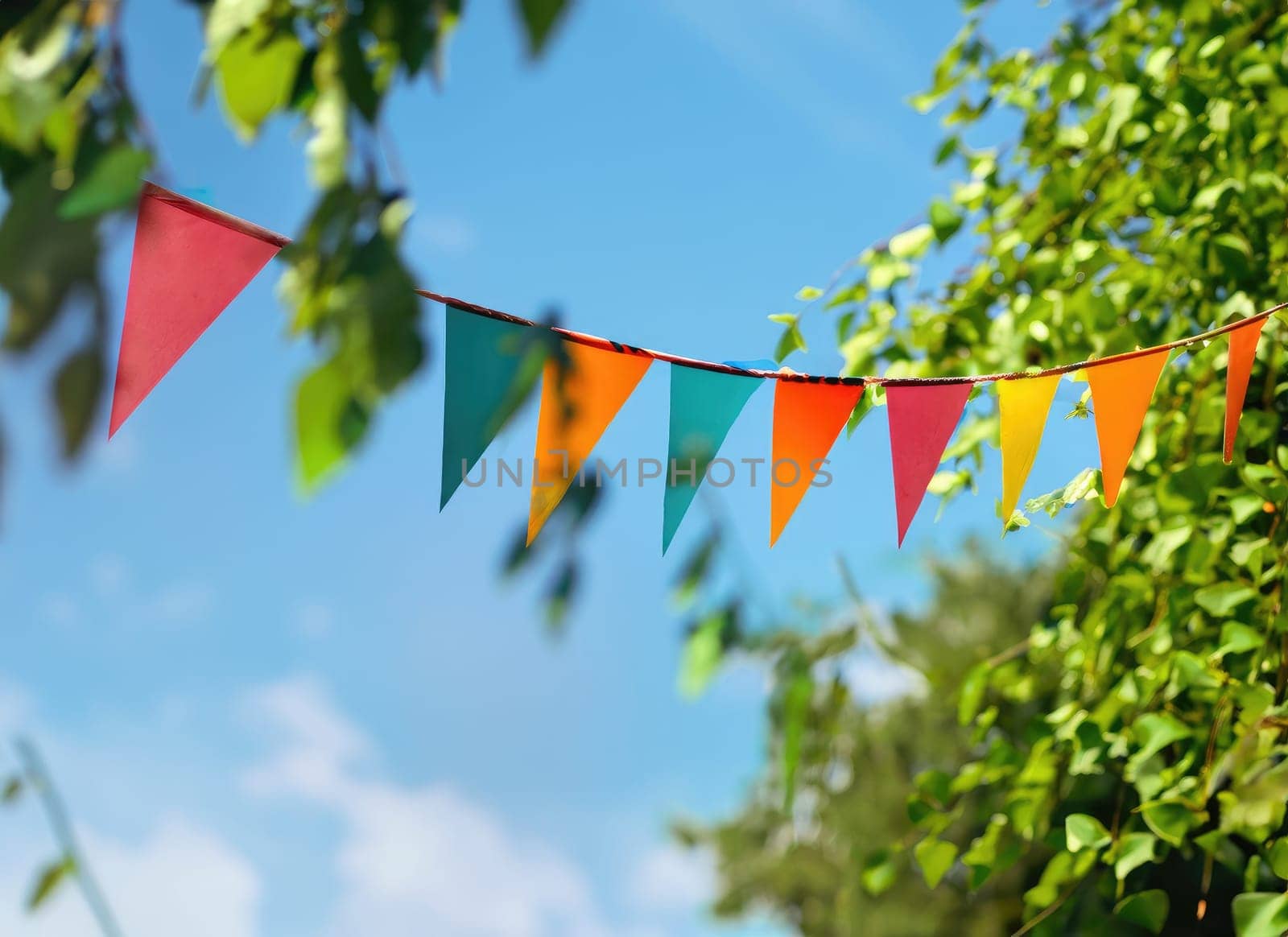 colorful pennant string decoration in green tree foliage on blue sky, summer party background.