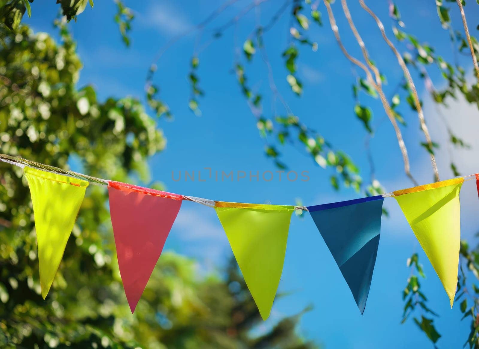 colorful pennant string decoration in green tree foliage on blue sky, summer party background.