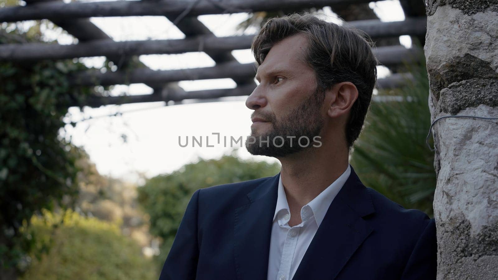 Portrait of a man looking in the distance in park. Action. Portrait of a male in blue suit standing outdoors, leaning on a stone wall