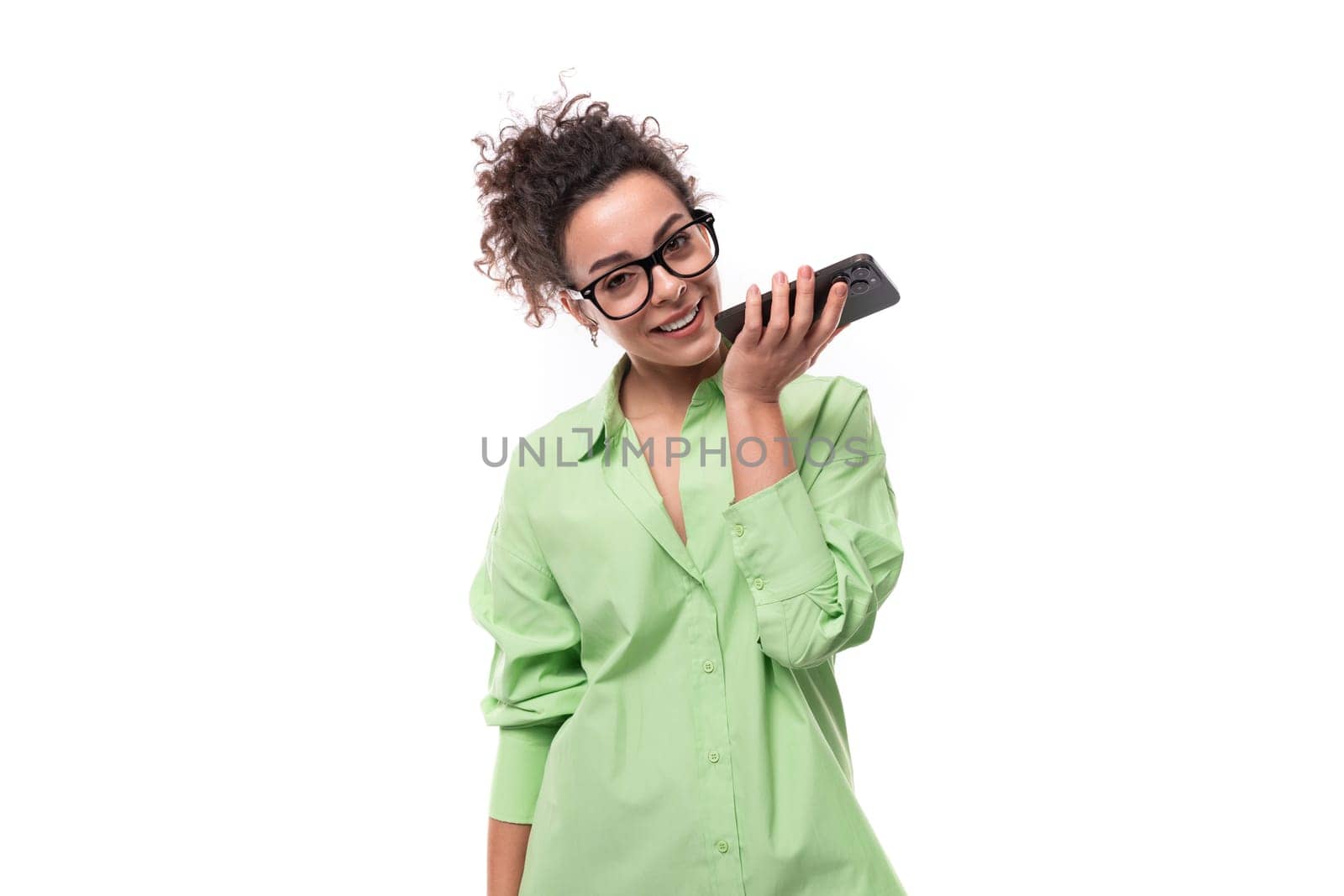 young stylish caucasian secretary woman with black curly hair wearing glasses and light green shirt is recording a voice message on the phone.