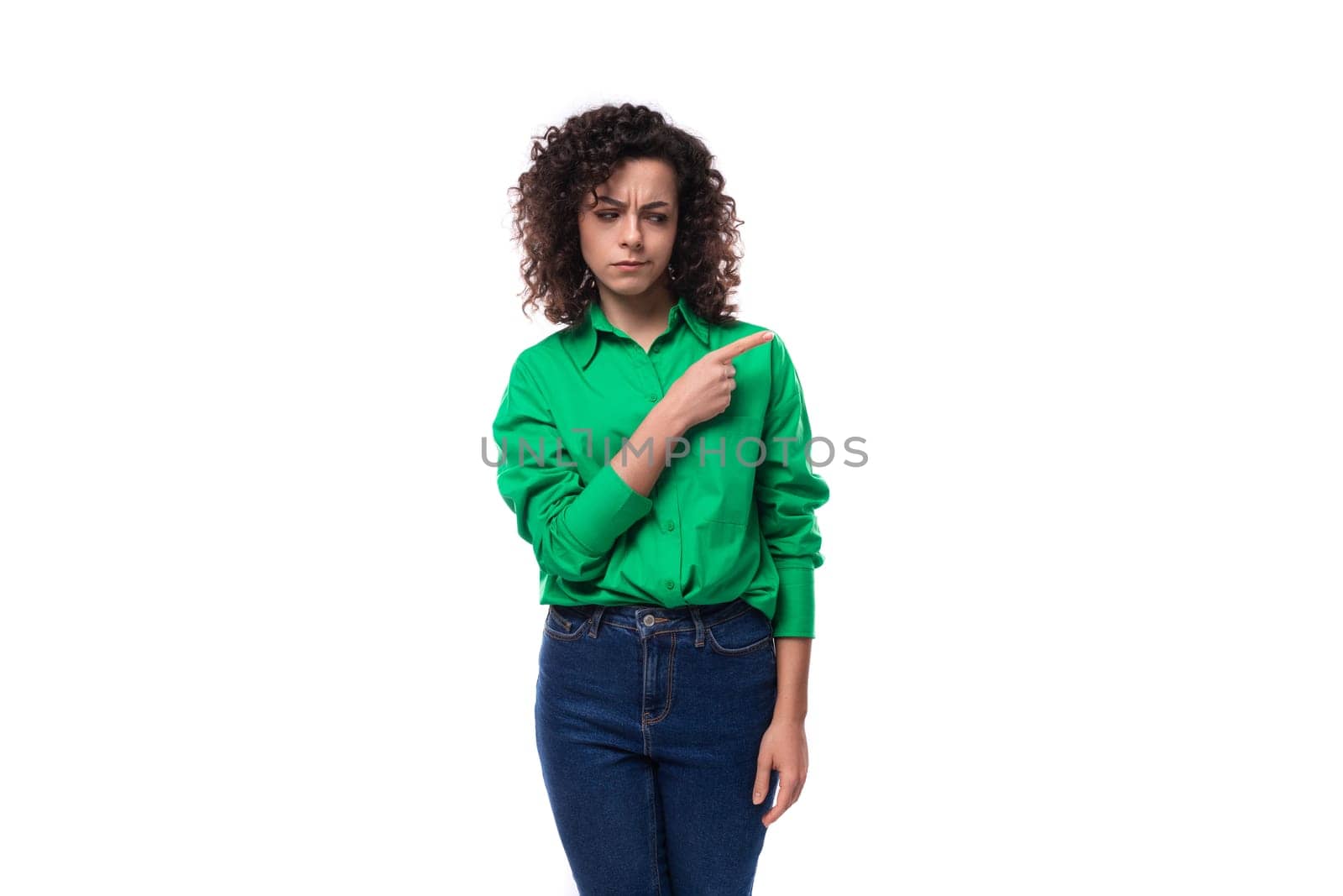 young caucasian brunette woman with curled hair dressed in a green shirt points her finger towards copy space. advertising concept.