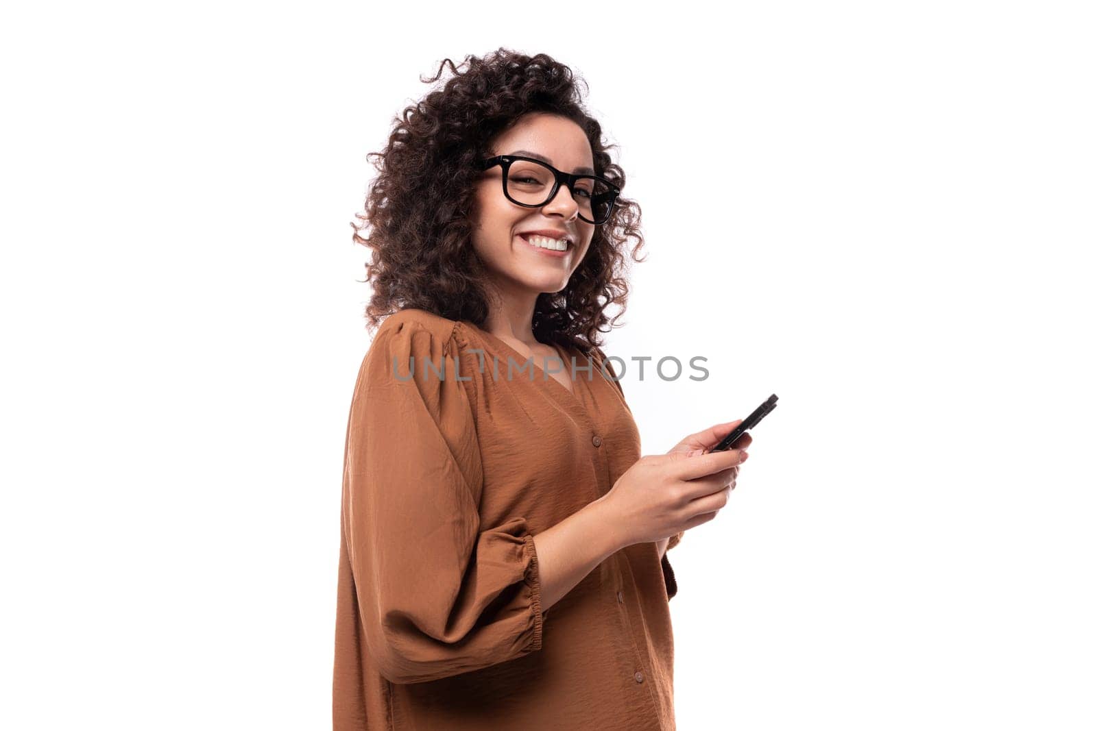young curly brunette woman dressed in a brown shirt chatting in a smartphone on a white background with copy space.