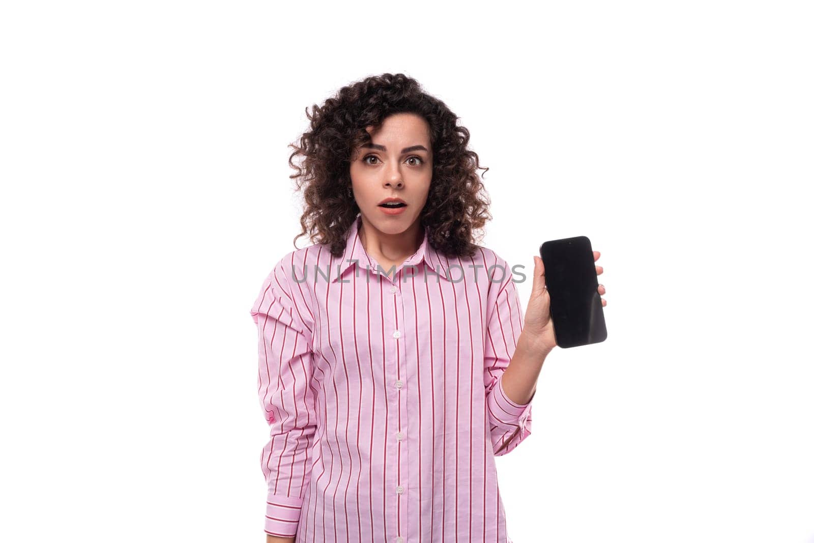 portrait of a surprised 30 year old slim curly brunette woman model dressed in a pink shirt with a smartphone in her hands. e-business concept.