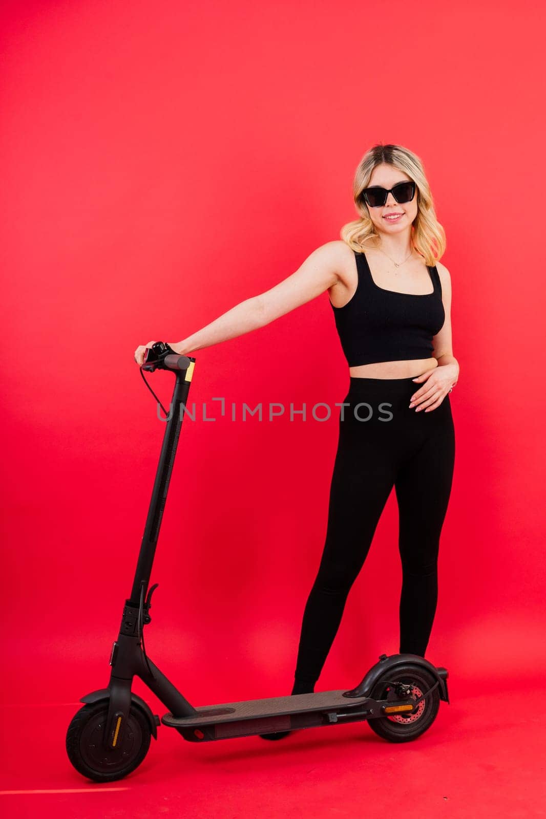 Beautiful emotional young female in sport clothes on electric scooter on red and white background by Zelenin