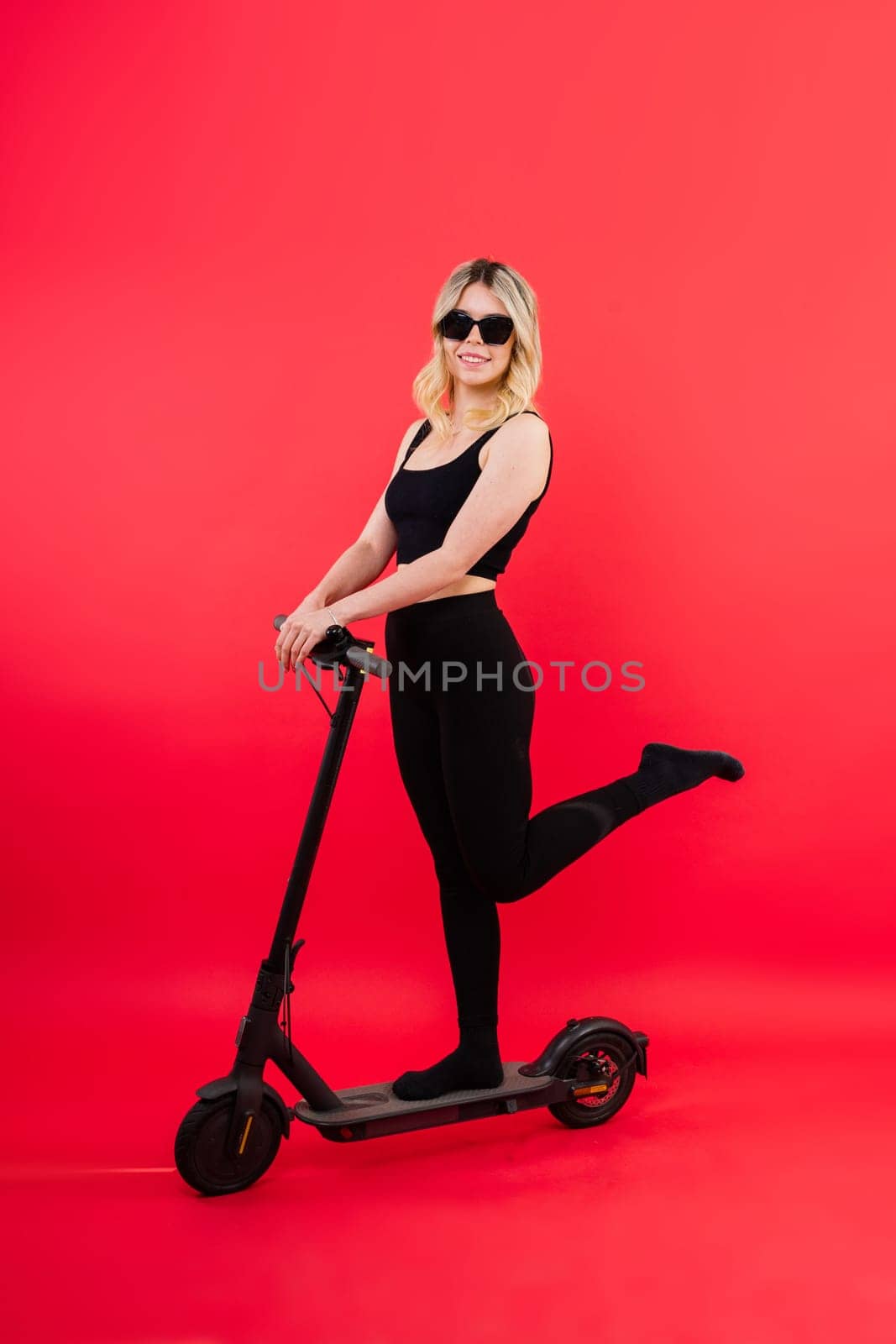 Beautiful emotional young female in a sport clothes on electric scooter on red and white background