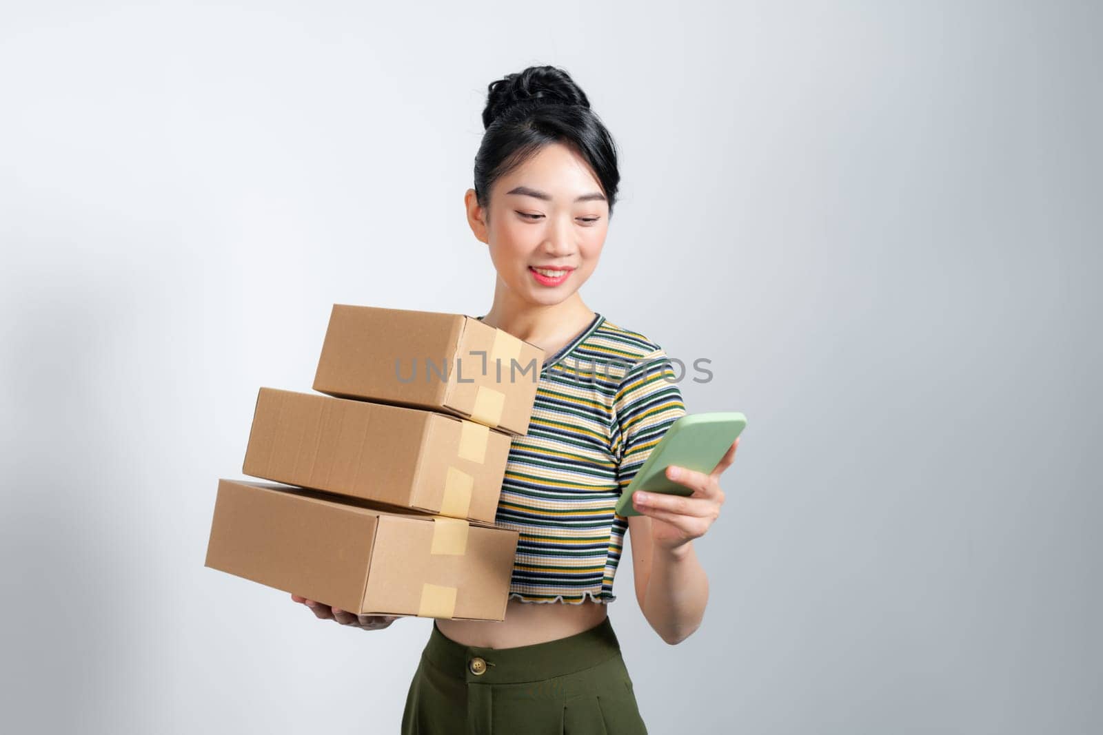 Young excited beautiful asian woman holding smartphone parcel cardboard standing on isolated white background.