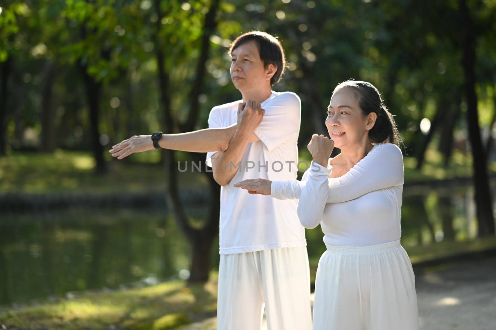 Happy retired couple in white clothes stretching in public park. Health care and wellbeing concept.