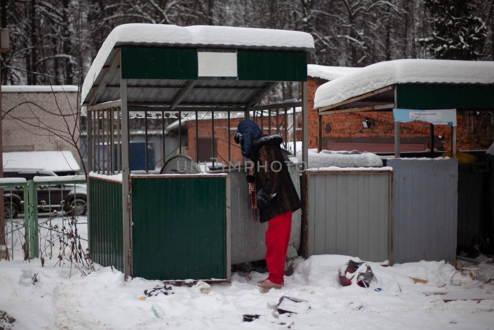 Man is looking for food in garbage. Man looks into dumpster in winter. Poor man on street. by OlegKopyov