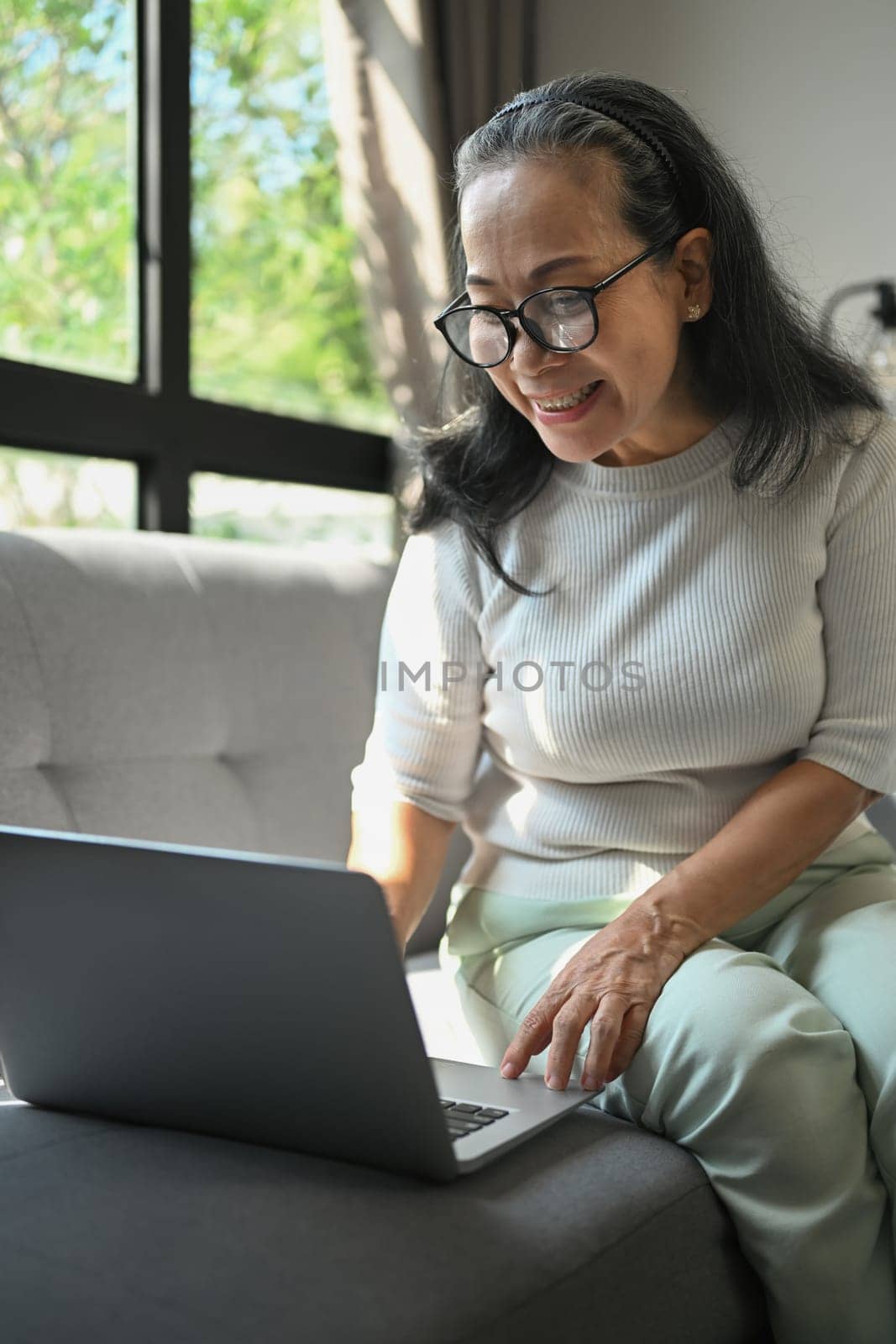 Happy middle age woman sitting on sofa chatting with friends in internet social networks.