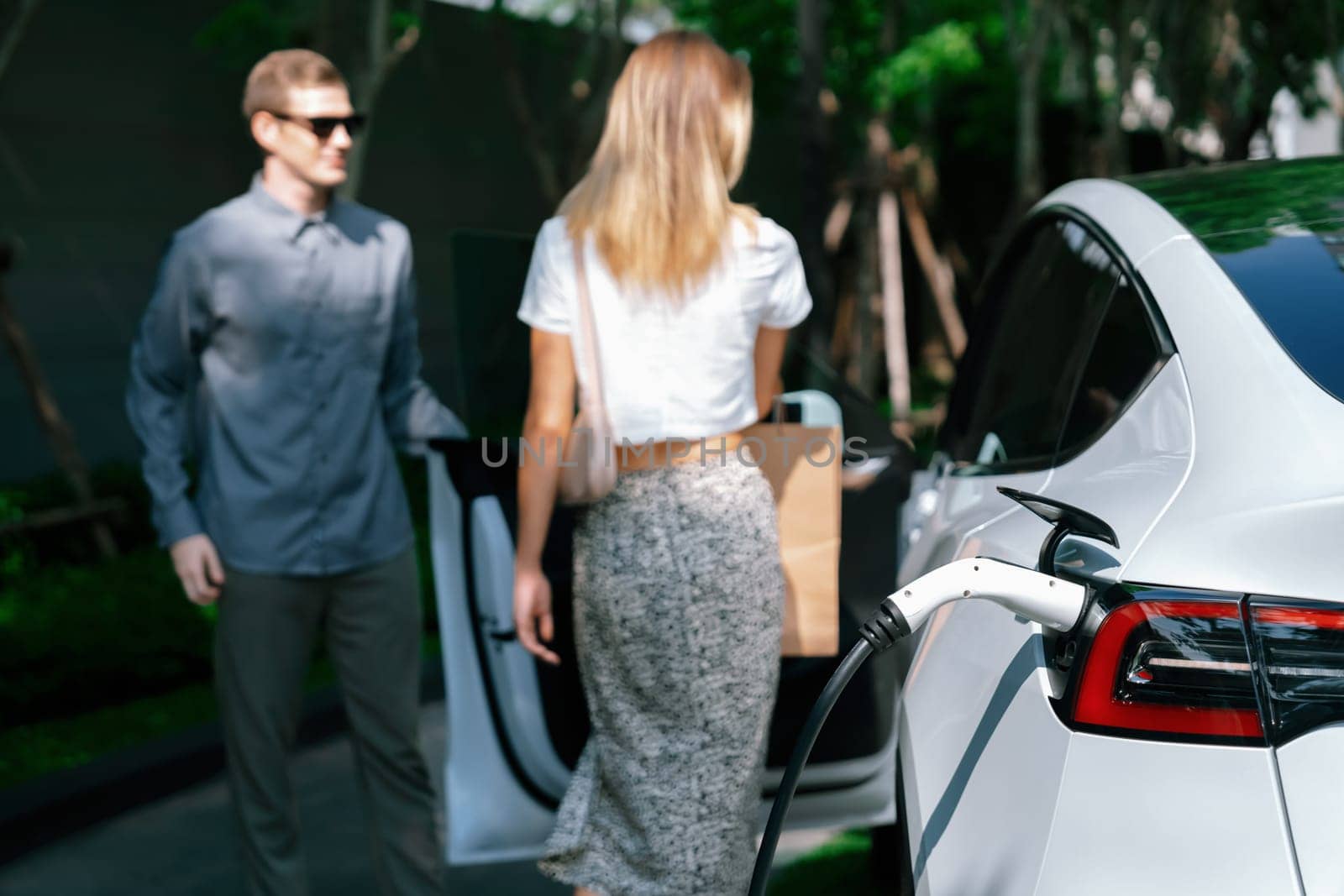 Young couple travel with EV electric car charging in green sustainable city outdoor garden in summer shows urban sustainability lifestyle by green clean rechargeable energy of electric vehicle innards