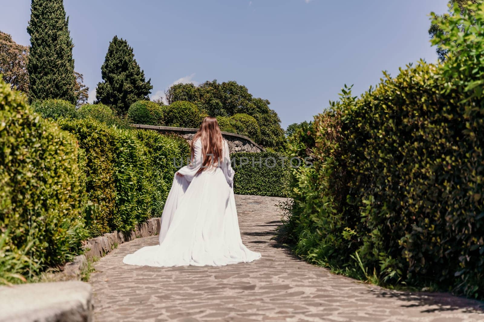 Brunette runs white dress park. A beautiful woman with long brown hair and a long white dress runs along the path along the beautiful bushes in the park, rear view.