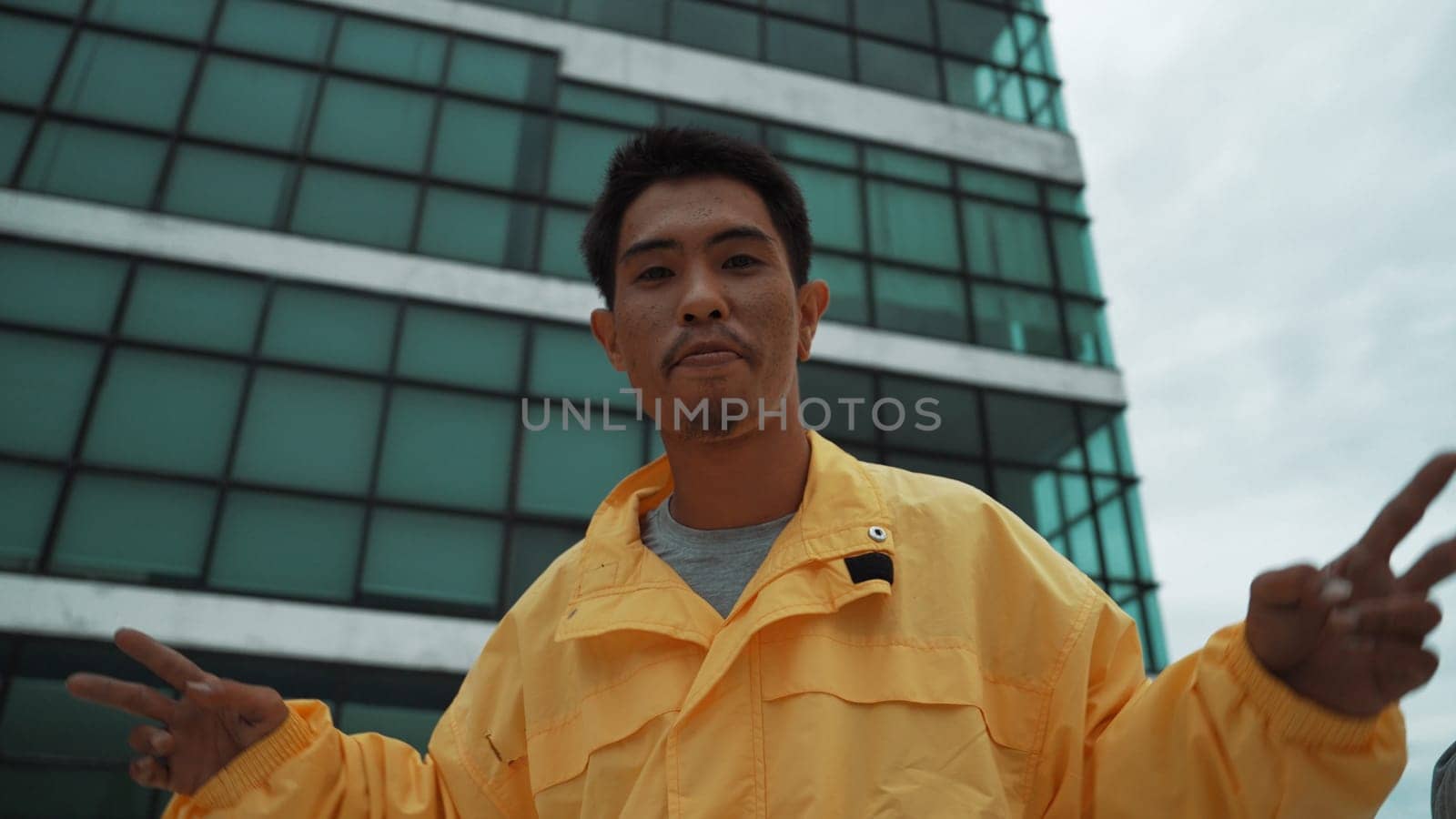 Hispanic street dancer posing at camera while standing at sky scrapper. Stylish attractive hipster moving his hands to hip hop music at rooftop with low angle camera. Outdoor sport 2024. Endeavor.
