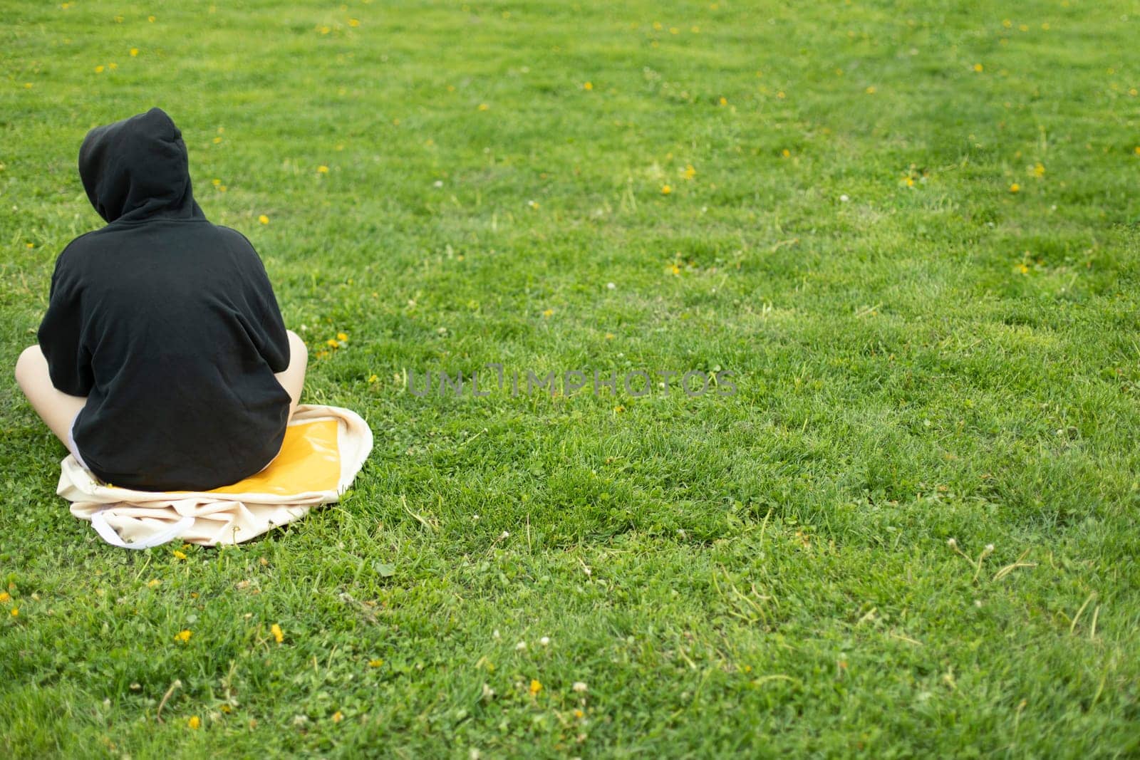 Teenager in black jacket in park. Girl in black hood sits on green grass. Alienated man. Teenage crisis.