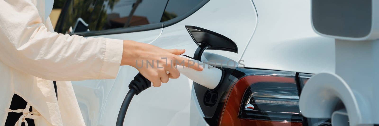 Young woman put EV charger to recharge electric car's battery from charging station. Alternative energy and rechargeable EV car for sustainable environmental friendly travel concept.Panorama Expedient