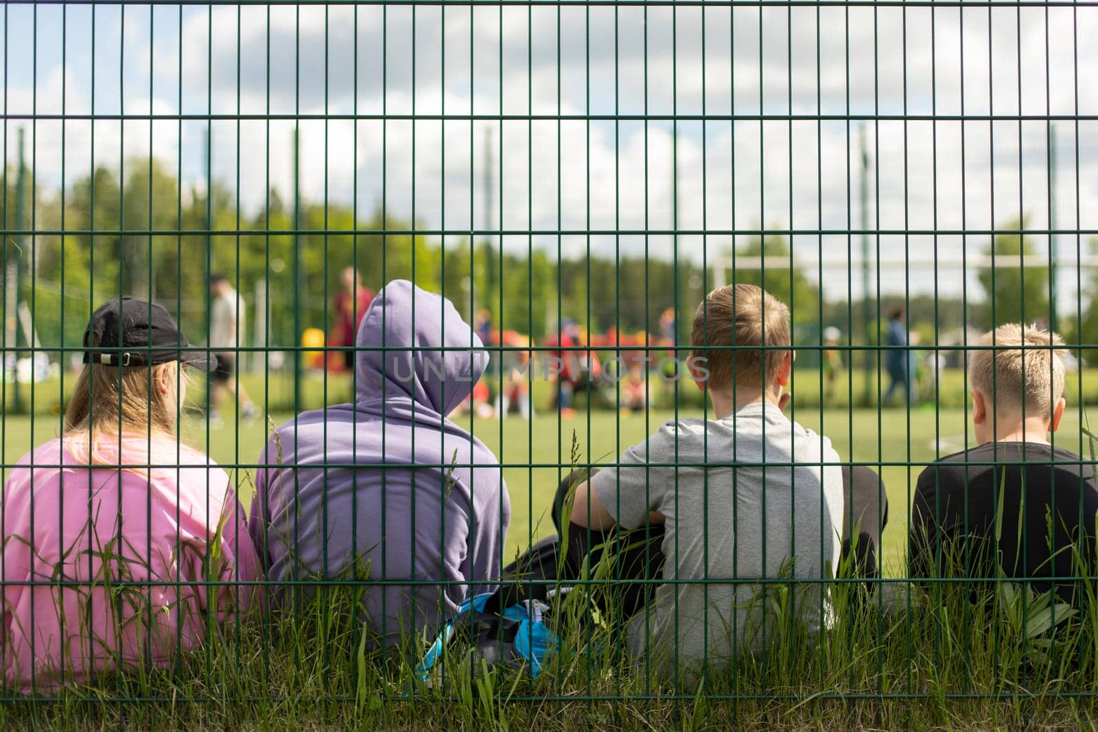 Children in stadium. Teenagers on sports field. Schoolchildren sit on grass behind net. Fence around football field. Spectators at sports competition.