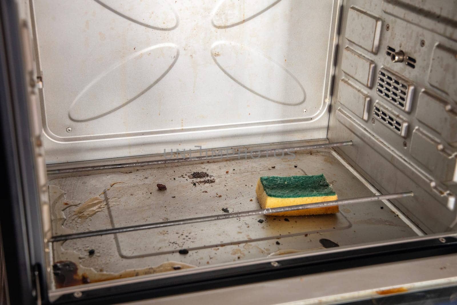 A yellow sponge with a cleaning solution lies in an open dirty oven with burnt grease stains on the internal surfaces, a freestanding oven on a table in the kitchen, high quality photo