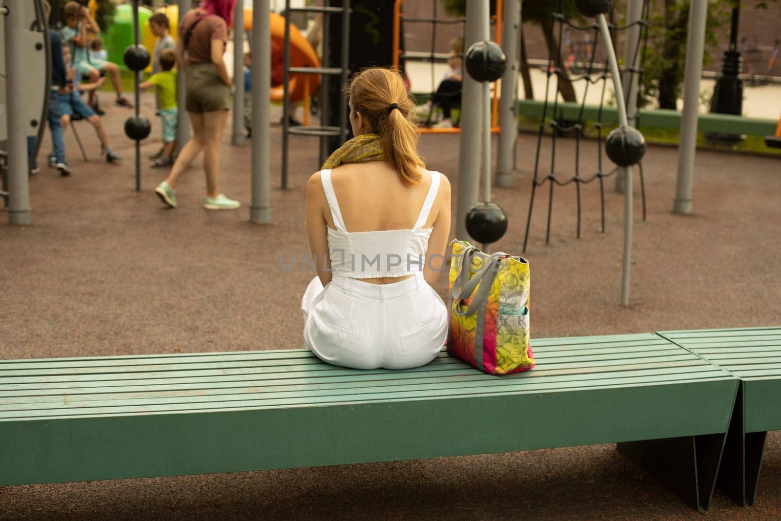 Girl on playground. Young mother watches her child in play area. Girl in white clothes.