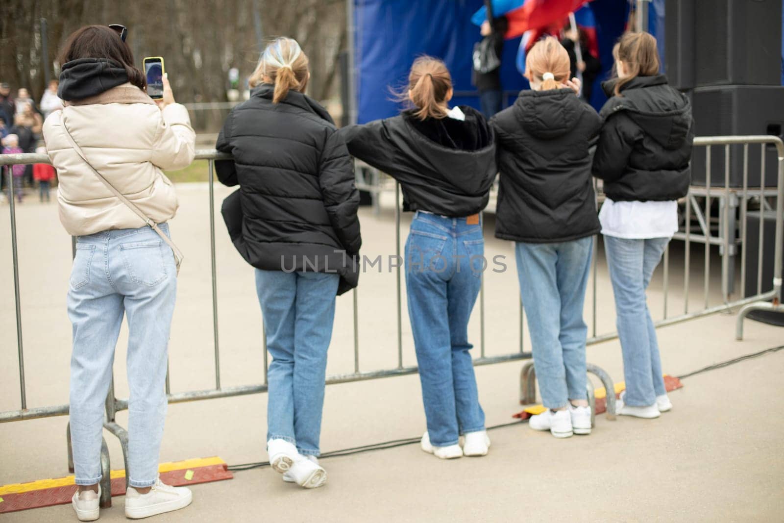 Girls stand at fence. Girls watch concert. People at city event. Girls in black jackets and blue jeans.