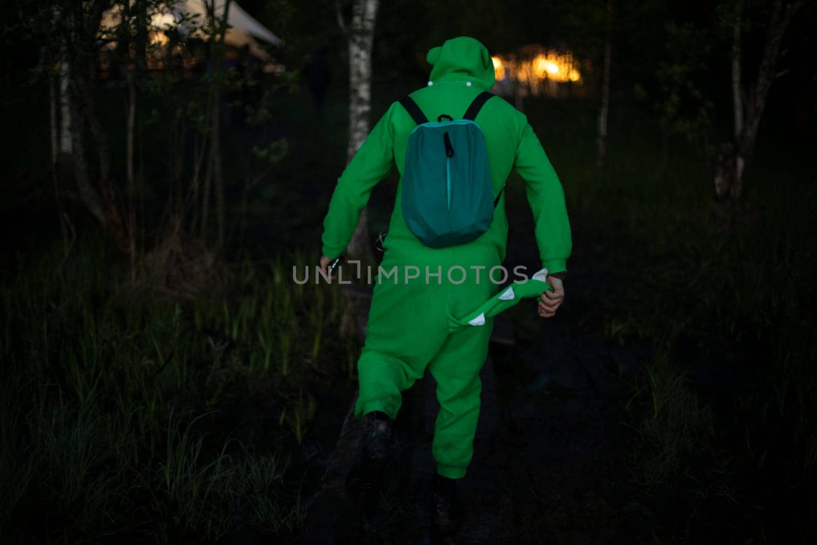 Man in green suit walks through woods. Guy in fun crocodile costume. Man at music festival in nature.