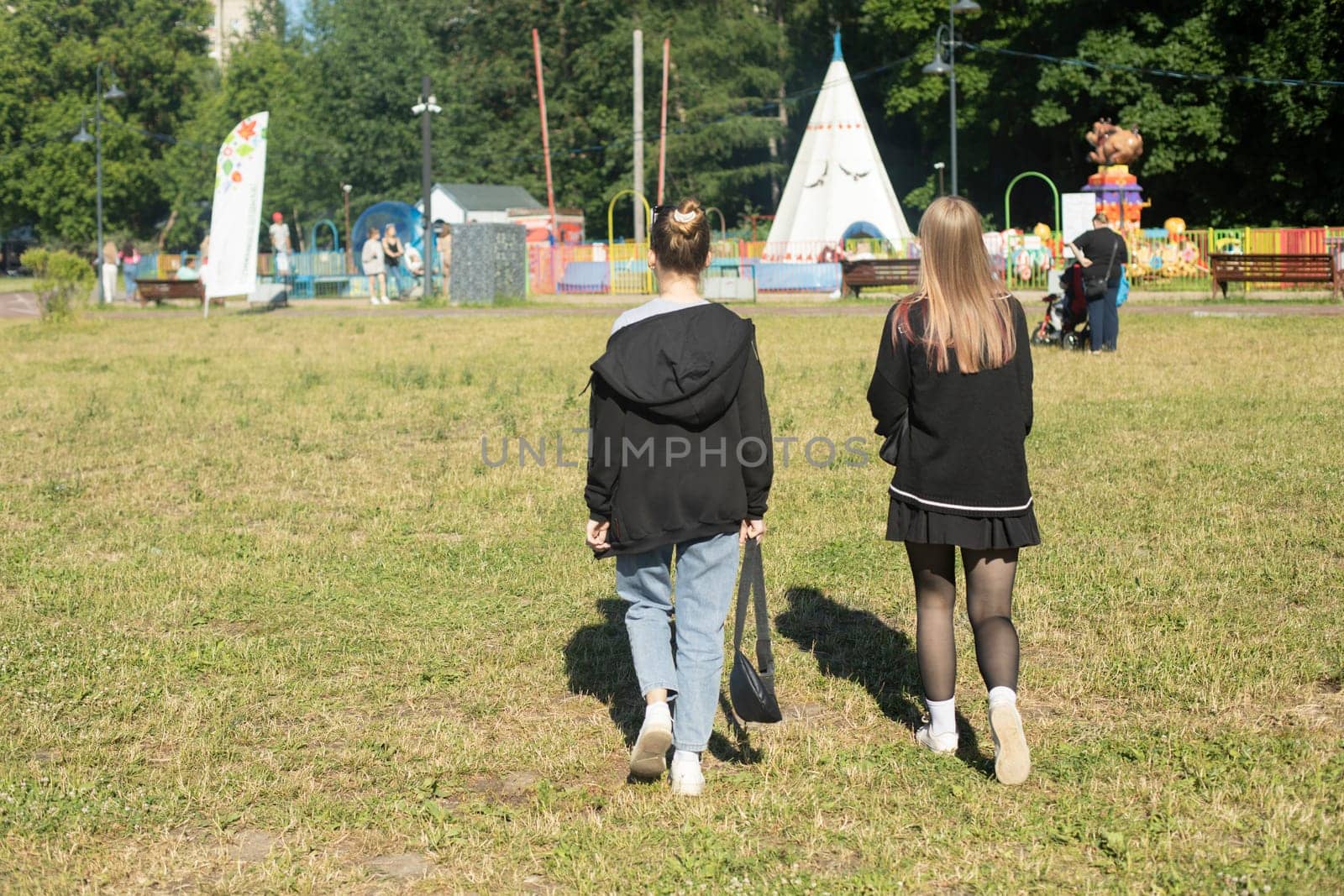 Girls walk in park in summer. Students at festival. Walking on green grass. Girls from back.