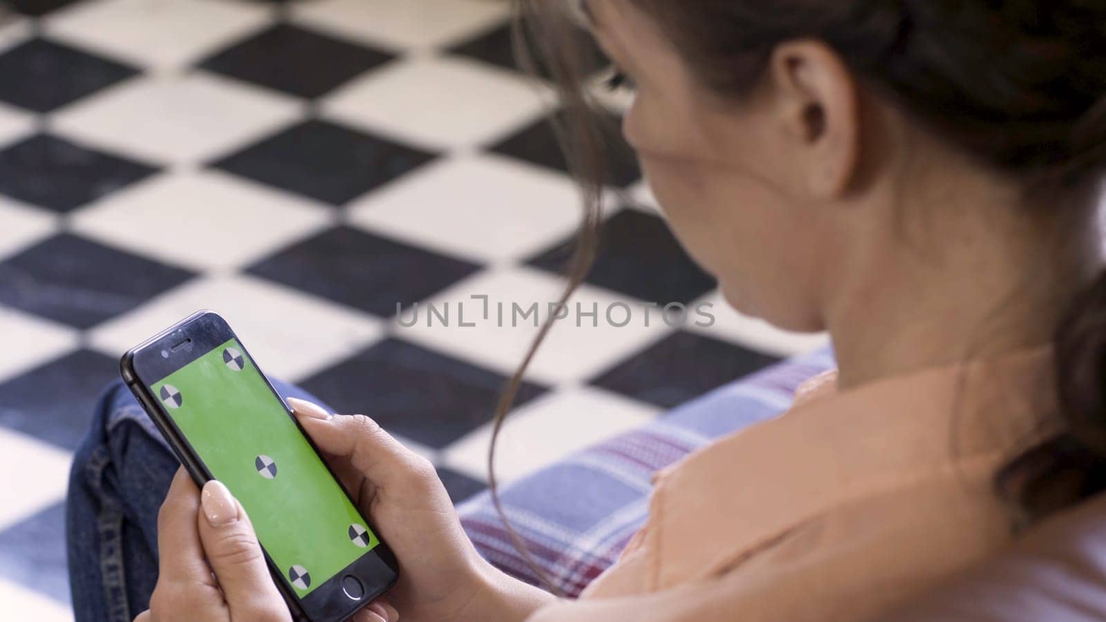 Beautiful woman holding a cell phone with green screen and tracking marks in her hands on checkered floor background. Woman holding mobile smart phone with chroma key.