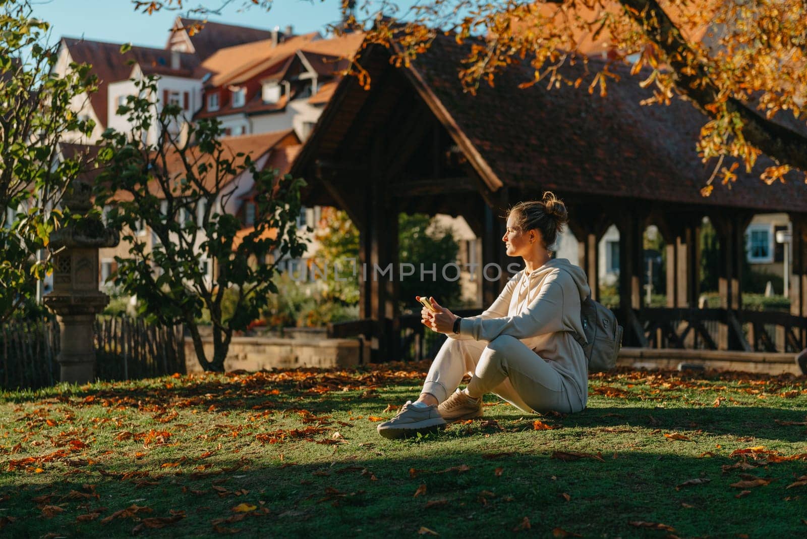 Young Fashionable Teenage Girl With Smartphone In Europian Park In Autumn Sitting At Smiling. Trendy Young Woman In Fall In Park Texting. Retouched, Vibrant Colors. Beautiful Blonde Teenage Girl Wearing Casual Modern Autumn Outfit Sitting In Park In Autumn. Retouched, Vibrant Colors, Brownish Tones. by Andrii_Ko