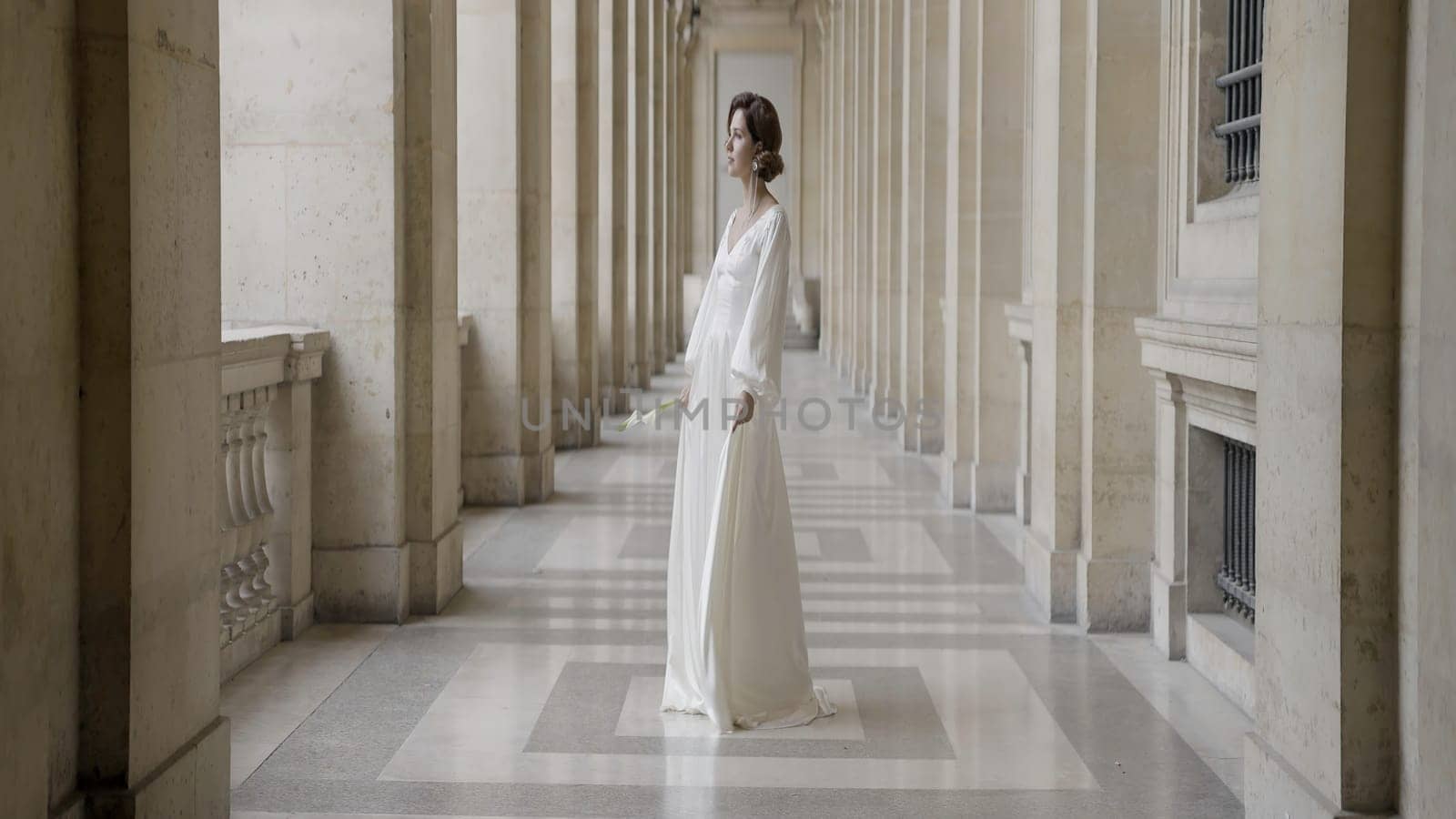 A beautiful model in a wedding dress and a white flower branch. Action. A young woman gently waving a long dress in different directions standing next to the architectural walls of the building. High quality 4k footage