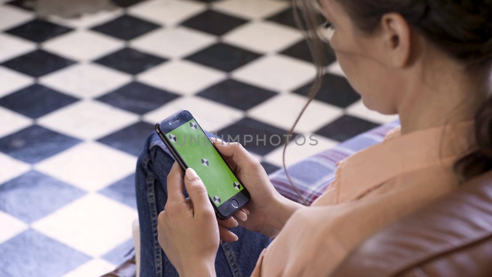 Beautiful woman holding a cell phone with green screen and tracking marks in her hands on checkered floor background. Woman holding mobile smart phone with chroma key.