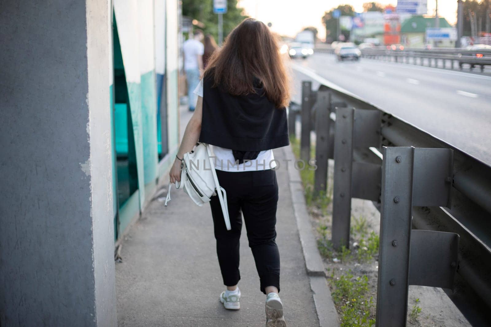 Girl walks along side of road. Girl in black jacket. Details of life in city.