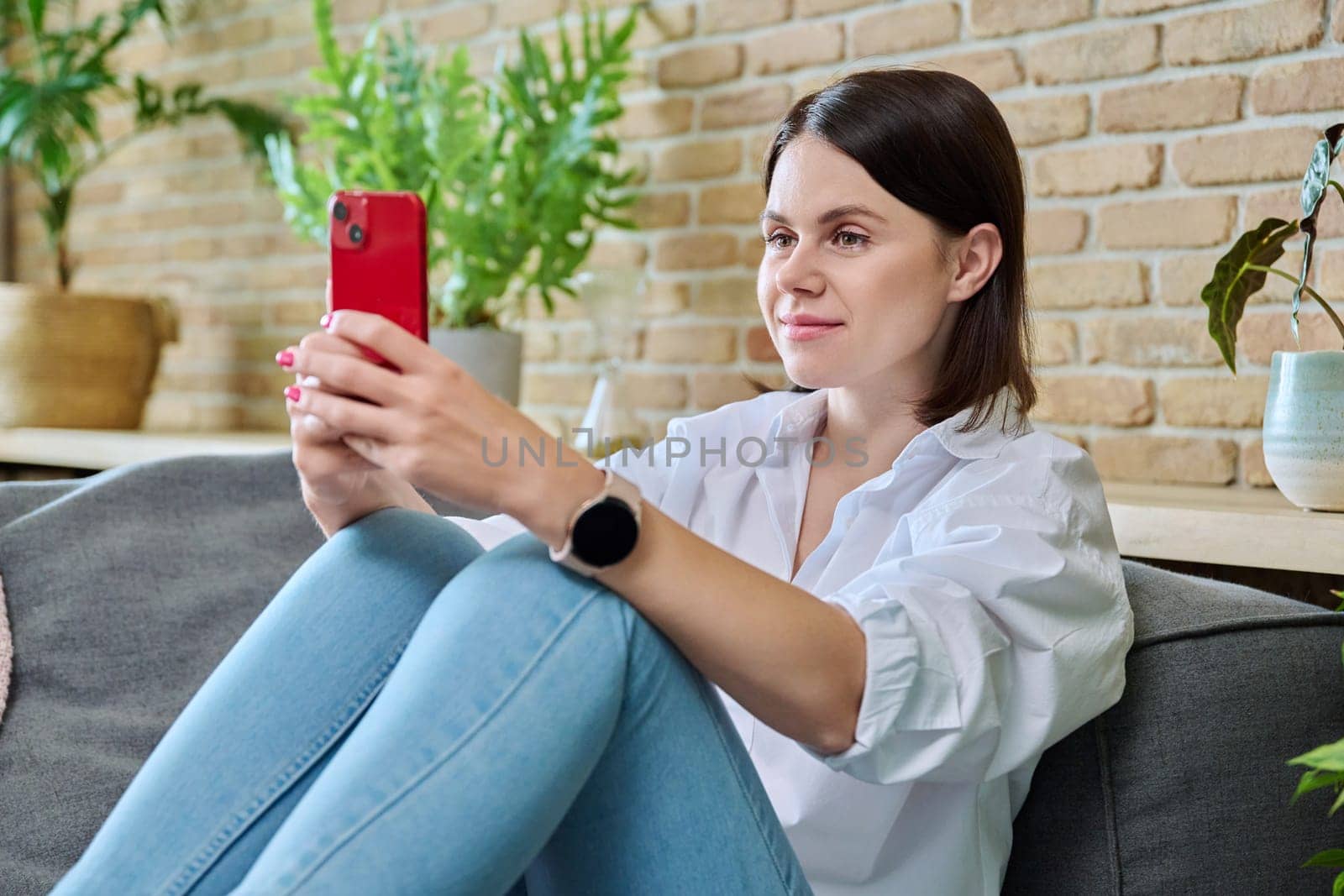 Young smiling happy woman using smartphone, sitting on sofa at home in living room. Internet online services, communication, mobile applications, technologies for leisure study work lifestyle