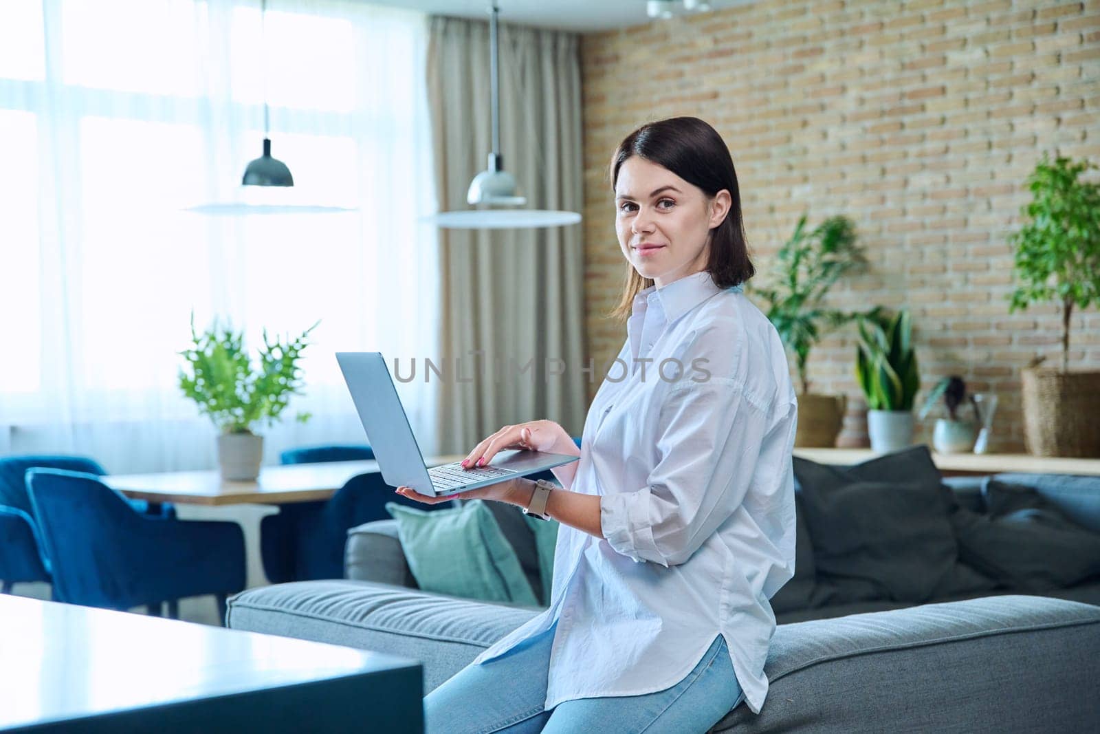 Young relaxed woman sitting on sofa at home in living room, looking at camera. Smiling female using laptop for work, freelancing, learning, blogging, communicating, typing on keyboard