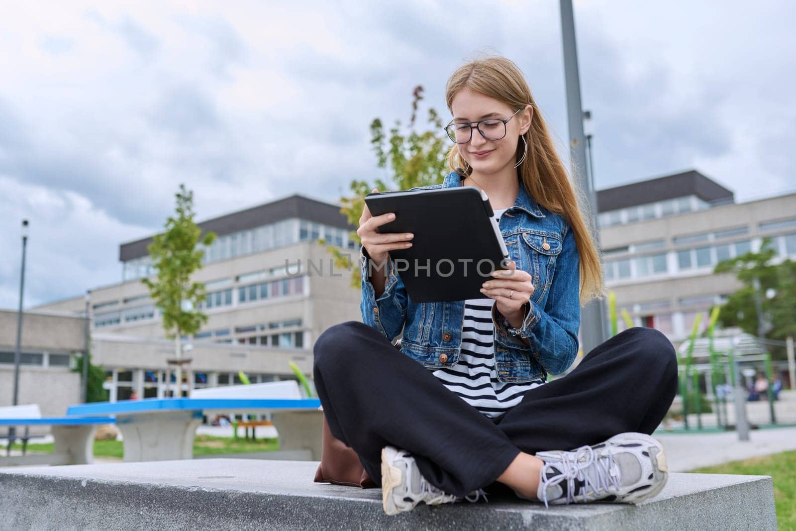 Girl student teenager outdoor near school building. Smiling teenage female with backpack, looking at screen of digital tablet. Adolescence, education, learning concept