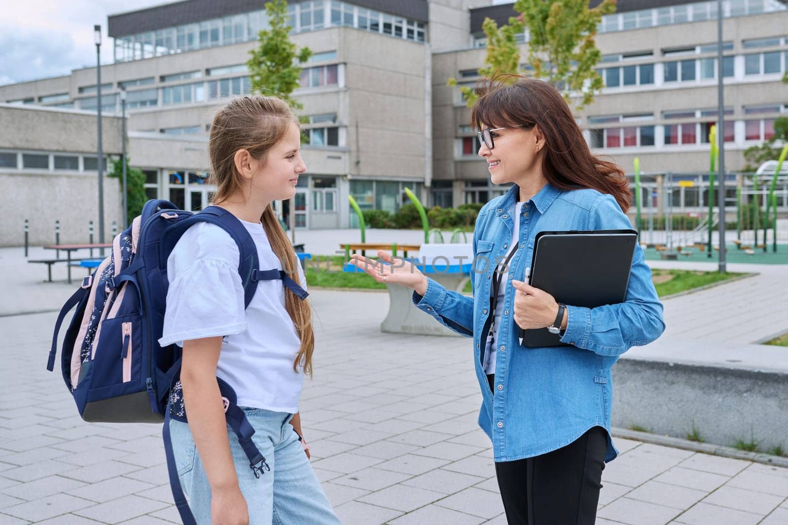 Talking female teacher and schoolgirl child outdoor, school building background. Meeting communication student girl with backpack and mentor counselor. Education, pre-teenage, learning, back to school
