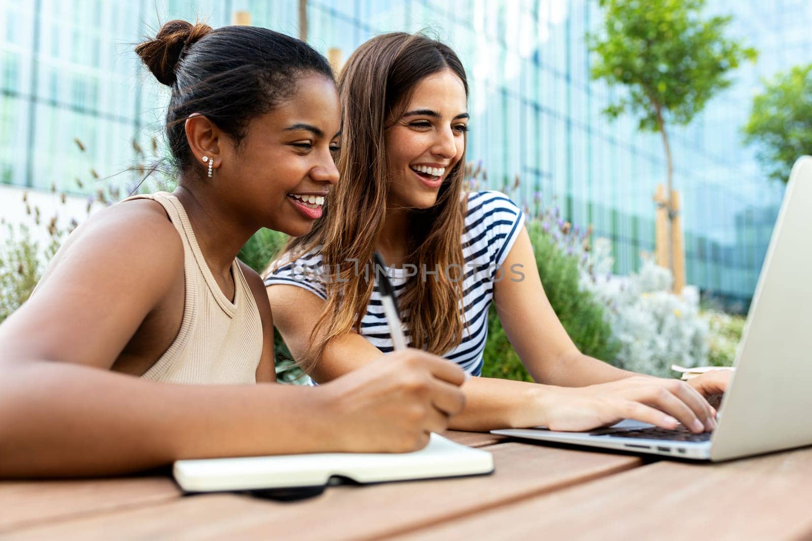 Two happy female college multiracial friends studying together in campus using laptop. Education and technology concept.