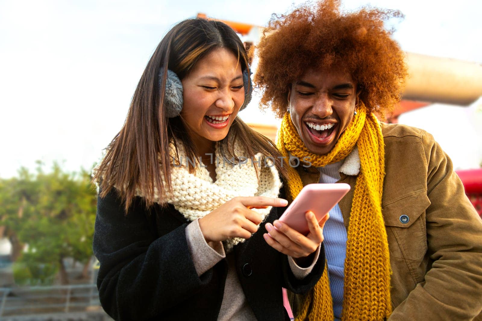Young adult multiracial couple having fun and laughing using a smartphone outdoors on a winter day. Cheerful multiethnic friends watching online videos on cellphone app. Two happy people using a mobile phone. Tehnology concept.