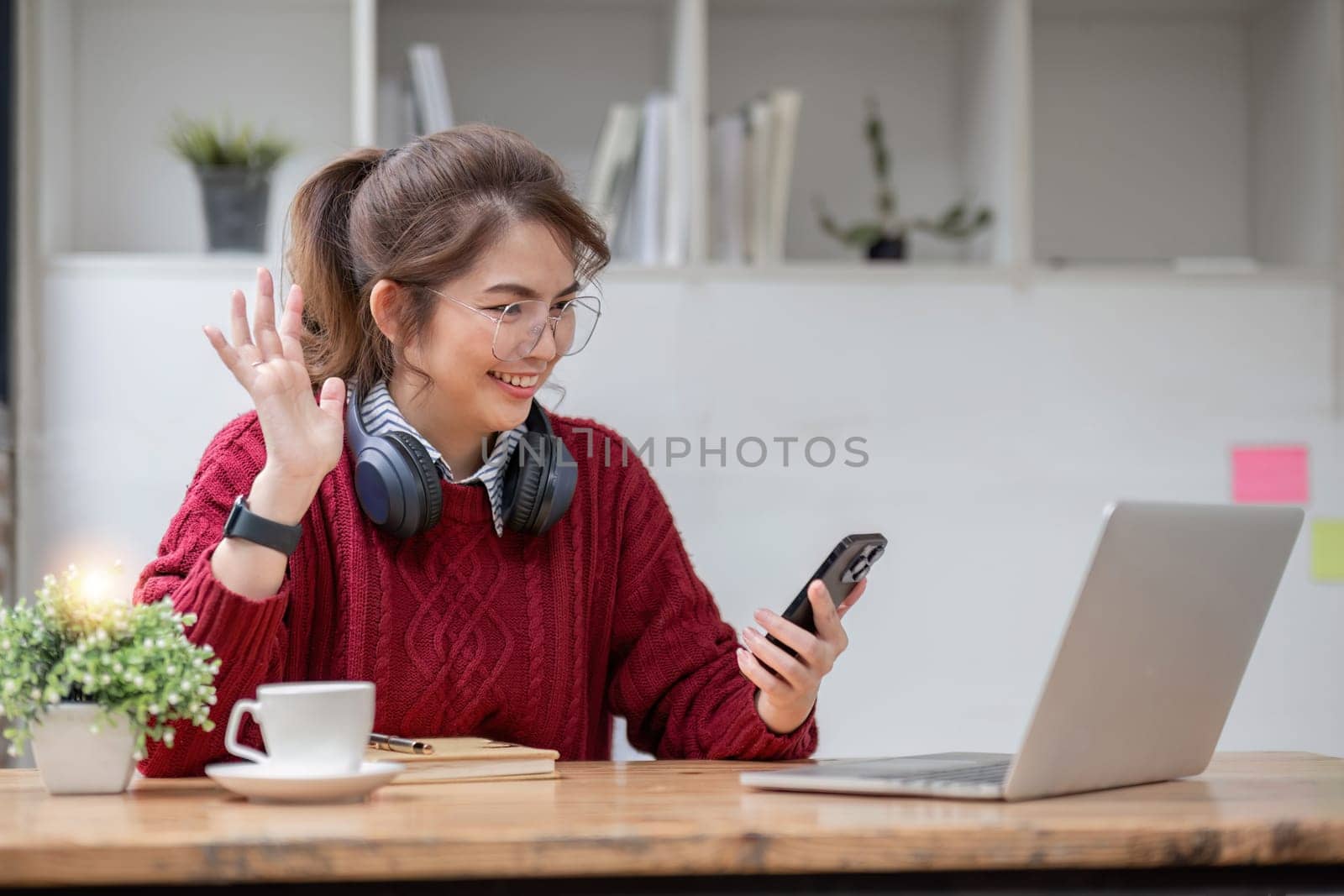 Asian female college student using laptop and phone with headphones while studying. Reading messages and greeting friends via video call by wichayada