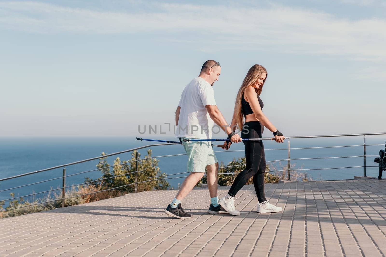 Happy Middle aged couple or friends practicing nordic walking in park near sea. Mature couple with trekking poles walking, practicing Nordic walking outdoors. Aging youthfully and sport concept.