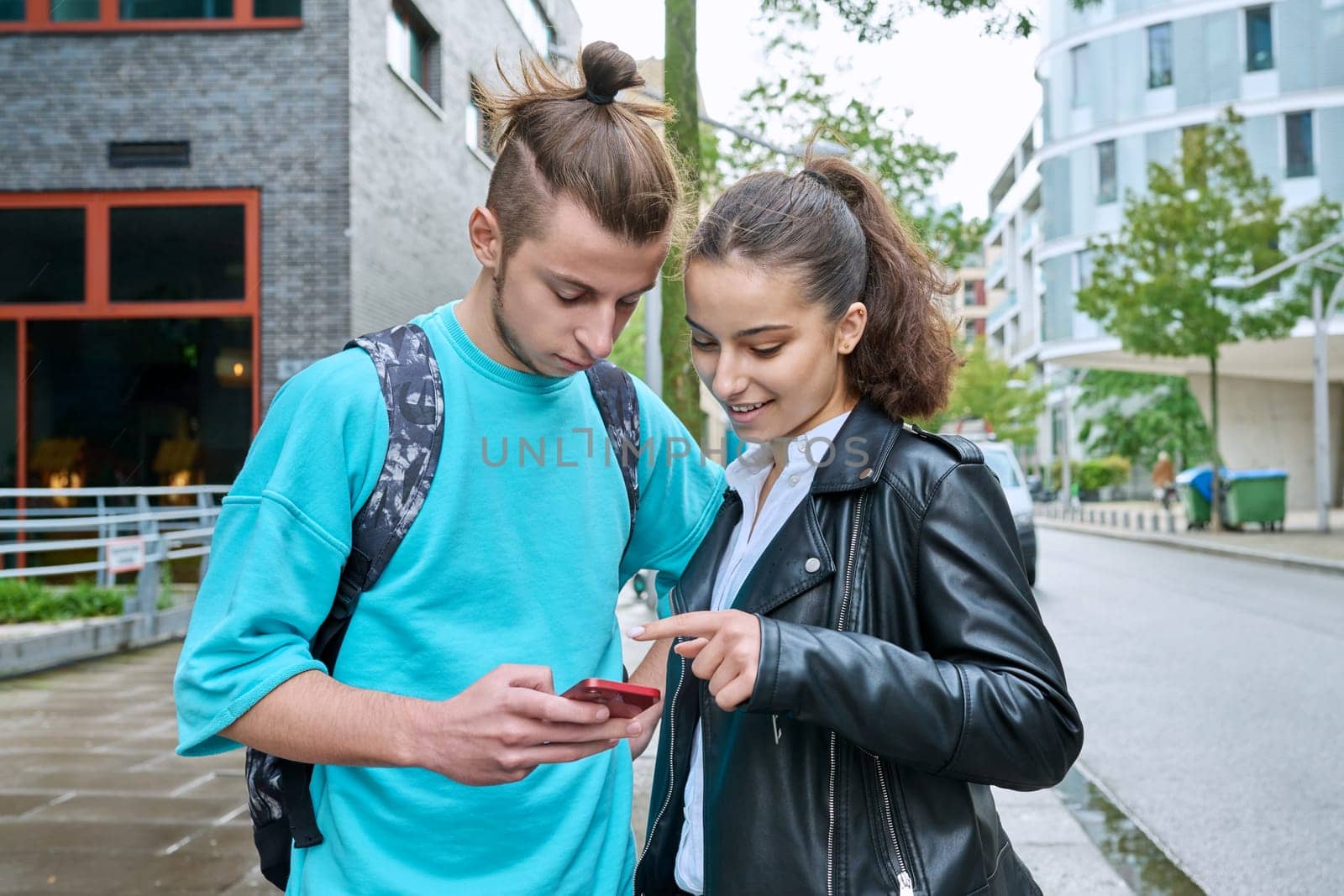 Teen friends guy and girl standing together holding smartphones looking at screen using mobile phones outdoor on city. Internet digital technology applications for leisure study communication