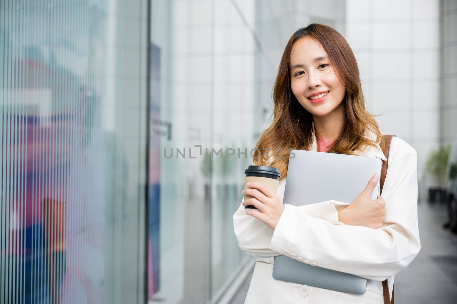 Beautiful portrait of a multitasking woman shows her walking against a beige wall with a takeaway coffee cup and a laptop in her hands. She smiling and confident, successfully work