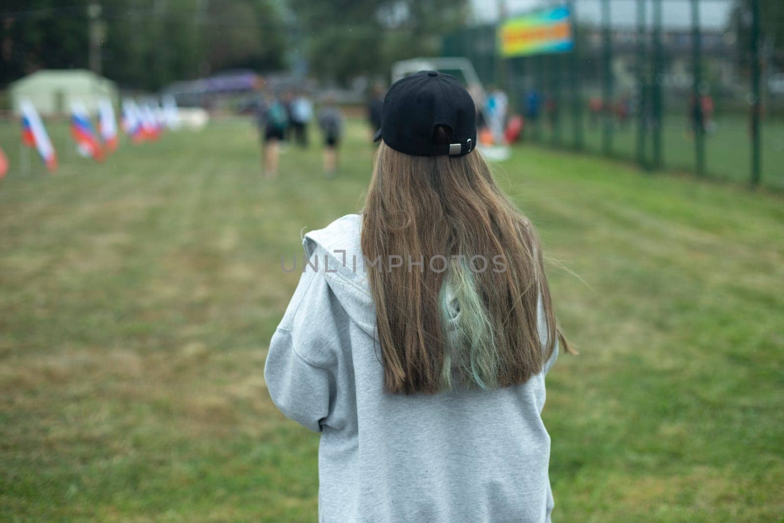 Girl in park. Teenager walks on street. Black cap and long hair. Grey jacket. Girl in summer.