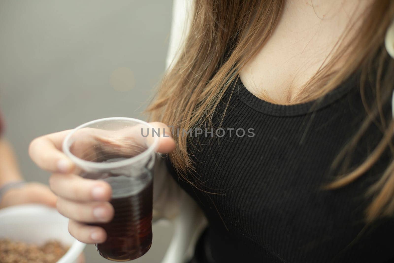 Girl holds glass with drink in her hand. Girl drinking tea outside. Black T-shirt and long hair. Hot drink in plastic cup.
