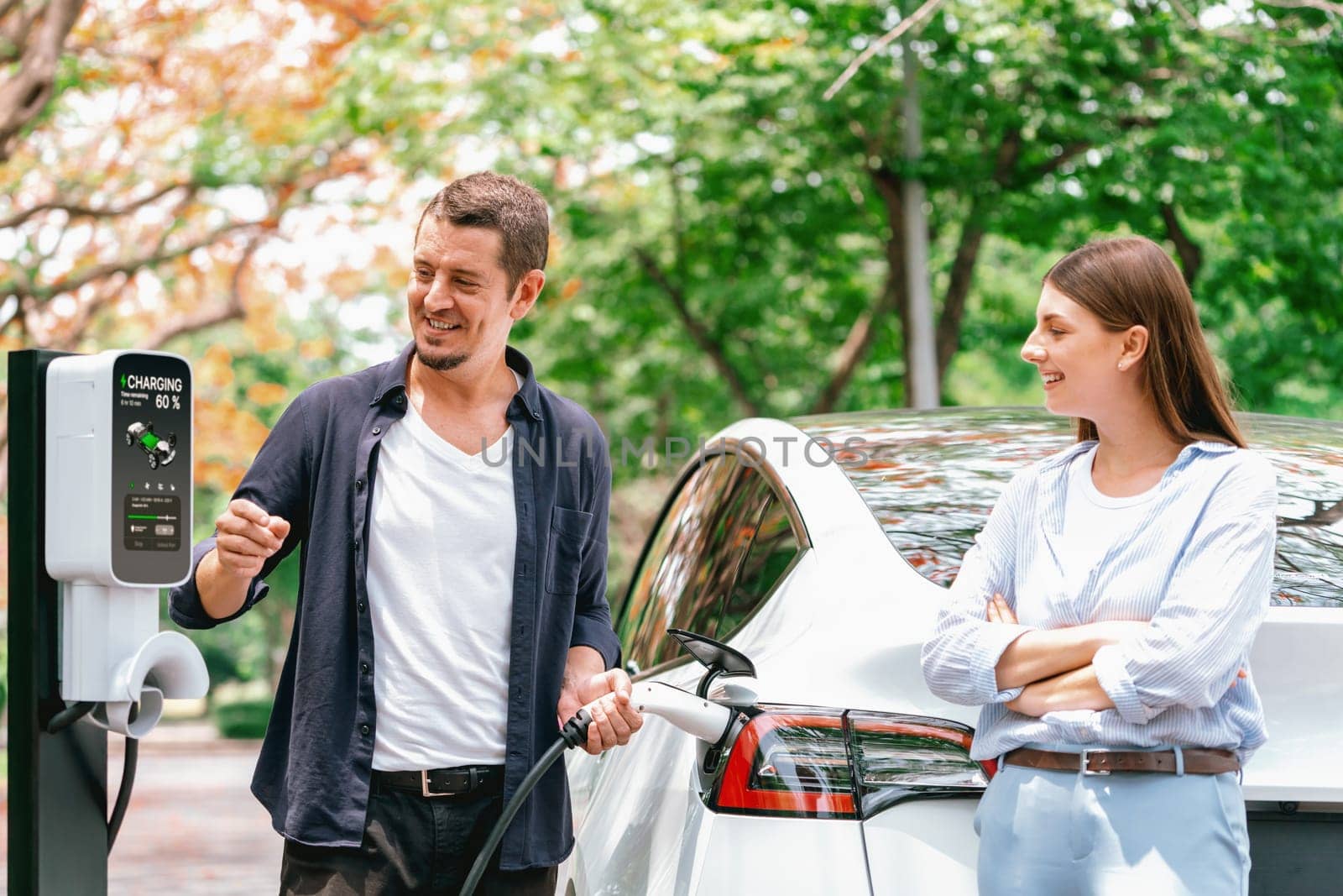 Lovely young couple recharging battery for electric car during autumnal road trip travel EV car in autumnal forest. Eco friendly travel on vacation during autumn. Exalt