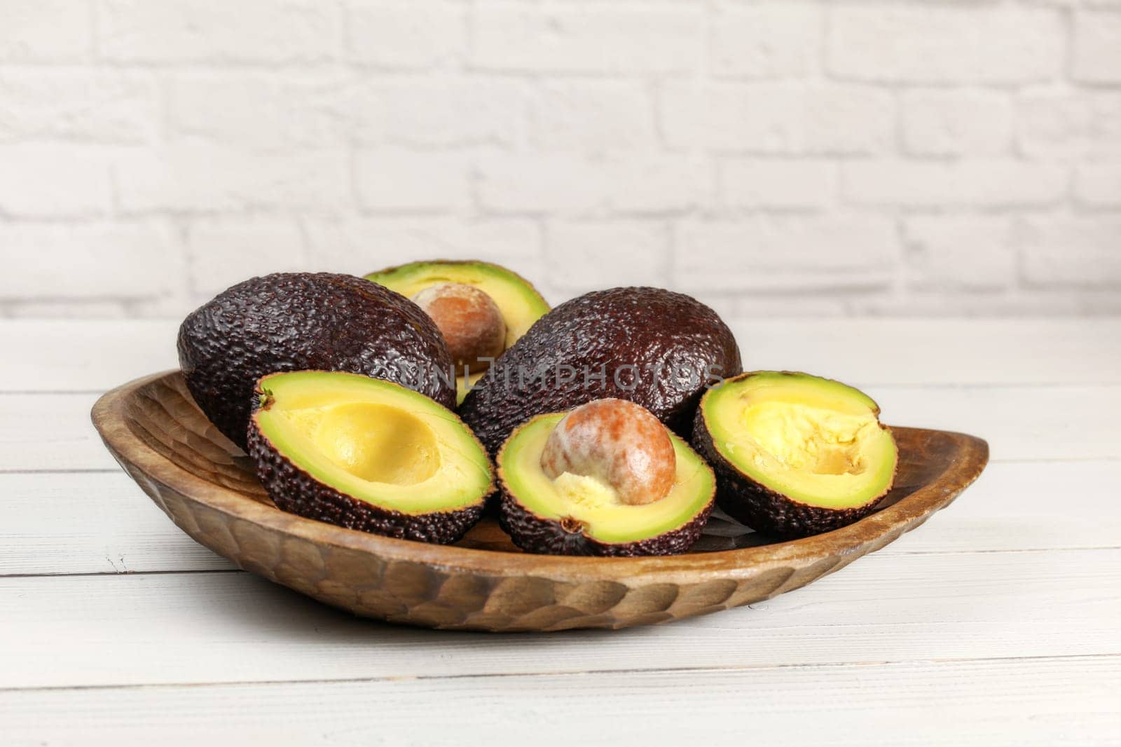 Ripe brown avocados ( hass - bilse variety ) some of them halved in wooden carved bowl, on white boards desk, brick wall background