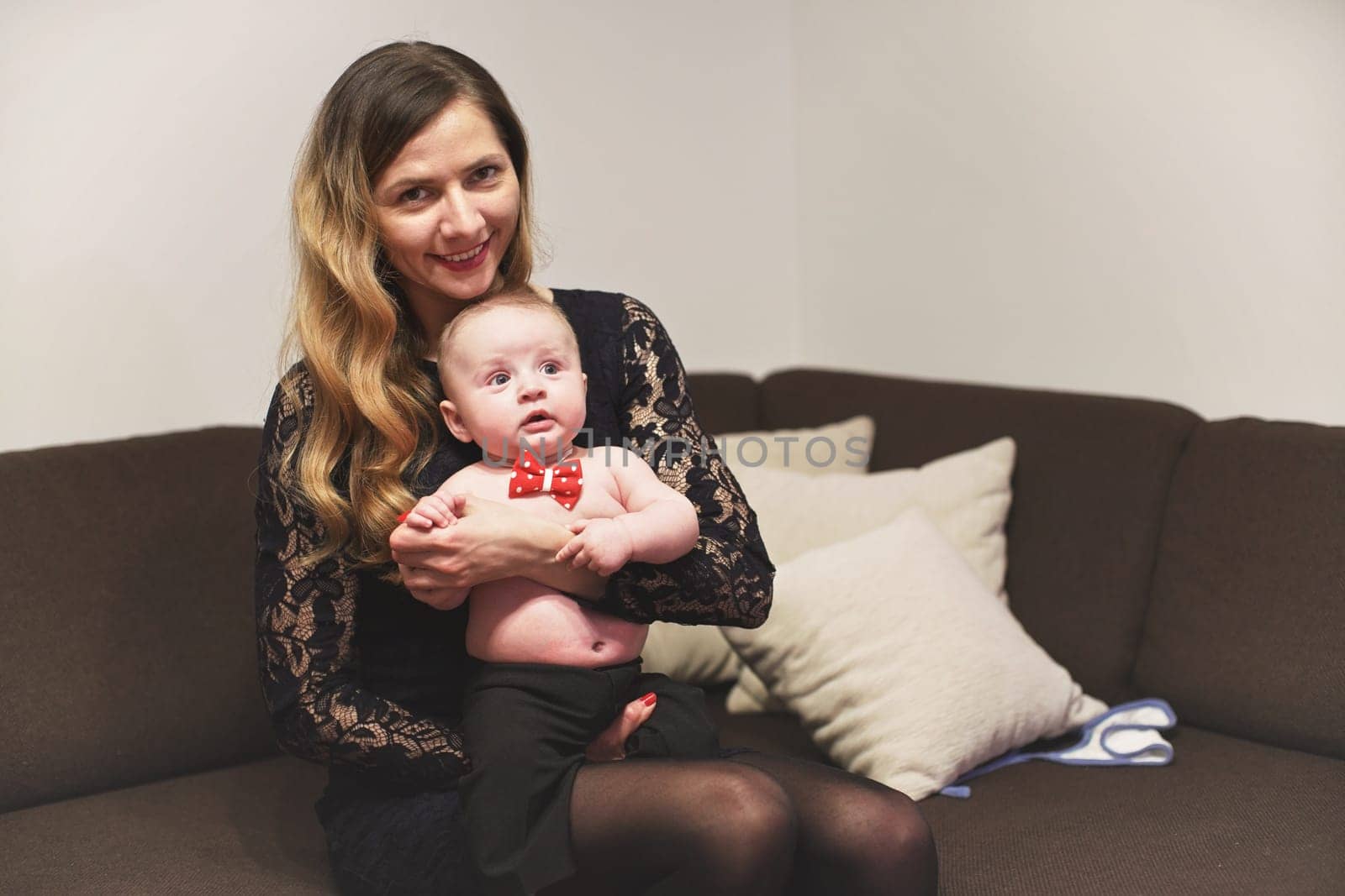 Young woman holding 4 months old infant baby boy with red bow tie on her hands, sitting on the couch