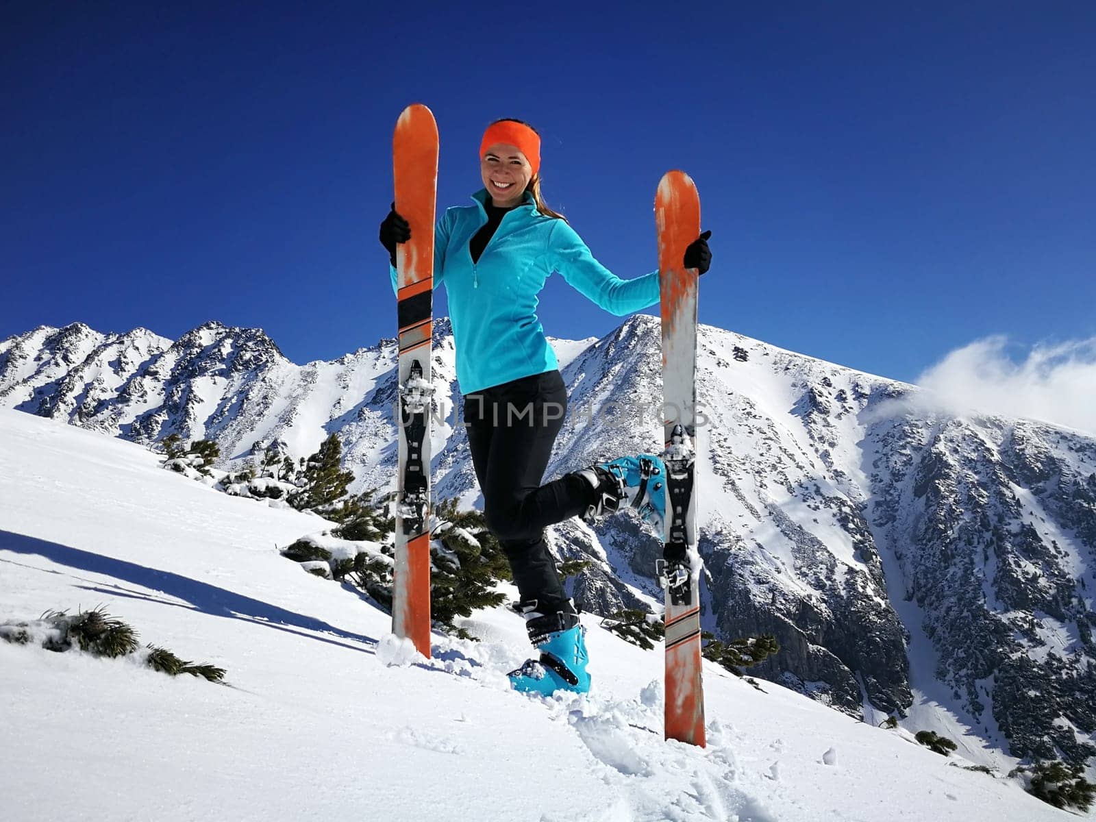 Young woman in bright blue - orange sport clothing and ski, posing, smiling happily on the hill, snow covered mountain, dark blue sky above, behind her by Ivanko