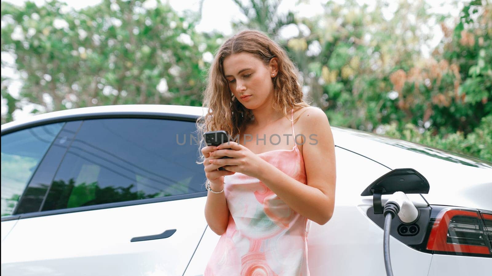 Young woman use smartphone to pay for electricity at public EV car charging station in nature. Modern environmental and sustainable automobile transportation lifestyle with EV vehicle. Synchronos